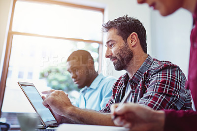 Buy stock photo Shot of man sitting at a table using a digital tablet with colleagues working around him