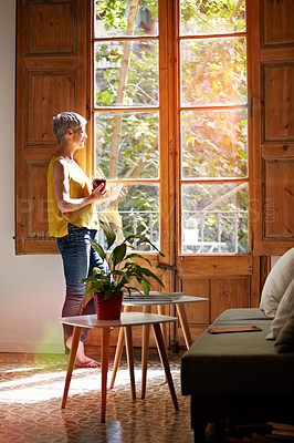 Buy stock photo Shot of a mature woman standing by a window at home drinking a cup of coffee