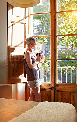 Buy stock photo Shot of a mature woman standing by a window at home drinking a cup of coffee