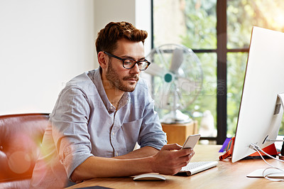 Buy stock photo Shot of a young man reading a text message while working on a computer in his home office