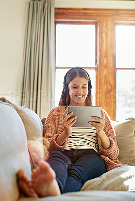 Buy stock photo Shot of a smiling young woman sitting on her sofa watching a movie on a digital tablet with headphones