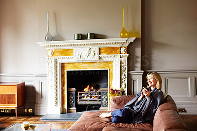 Buy stock photo Shot of a mature woman sitting on her sofa drinking tea while wrapped in a blanket