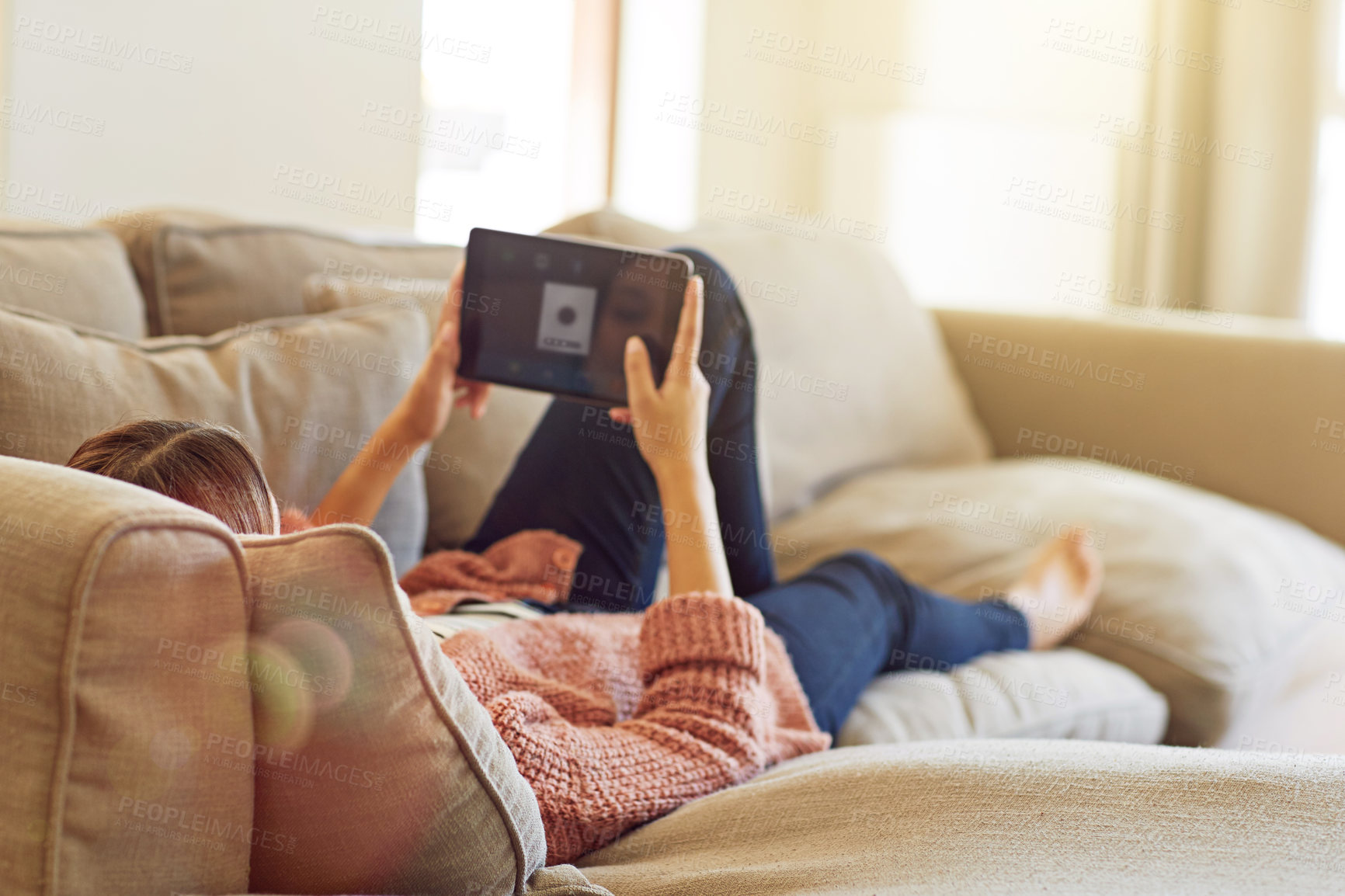 Buy stock photo Rearview shot of a young woman lying on her sofa watching a movie on a digital tablet