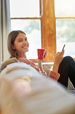 Buy stock photo Portrait of a young woman sitting on her living room sofa drinking a coffee and using a cellphone