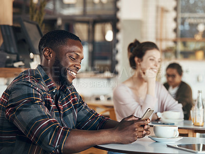 Buy stock photo Shot of a handsome young man sitting in a busy coffee shop while using his cellphone during the day