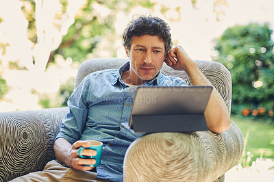 Buy stock photo Shot of a mature man relaxing in a chair while using a digital tablet and drinking coffee in his backyard