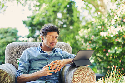 Buy stock photo Shot of a mature man relaxing in a chair while using a digital tablet and drinking coffee in his backyard