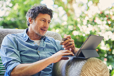 Buy stock photo Shot of a mature man relaxing in a chair while using a digital tablet and credit card in his backyard
