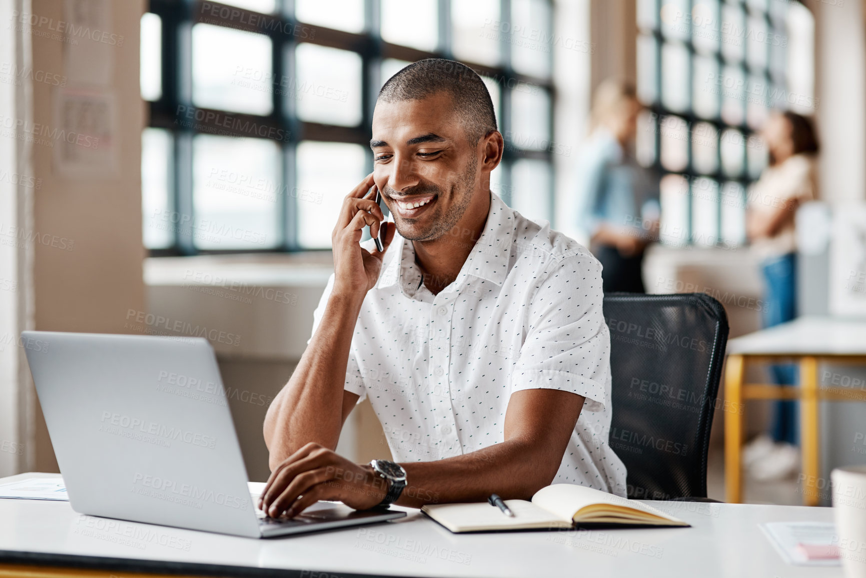 Buy stock photo Shot of a young businessman talking on a cellphone while working on a laptop in an office