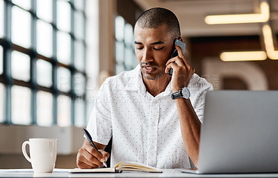 Buy stock photo Shot of a young businessman using a laptop, smartphone and making notes in a modern office