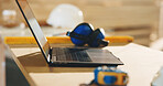 Carpentry, laptop and earmuffs on desk n workshop for hearing protection or safety equipment. Woodworking, computer and noise reduction headphones on table for timber project in empty factory.