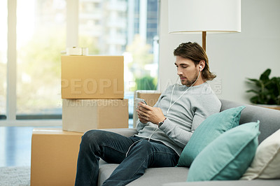 Buy stock photo Man, phone and relax on sofa with moving, earphones and listen to music, texting and web chat in new home. Person, smartphone and scroll with mobile app, streaming subscription and boxes at apartment