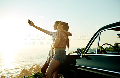 Buy stock photo Shot of two young women taking selfies with a mobile phone on a road trip