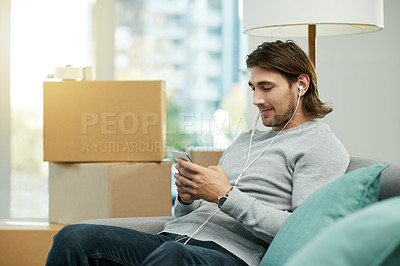 Buy stock photo Man, phone and boxes in new home, sofa and listen to music with earphones, texting or web chat in lounge. Person, smartphone and moving with mobile app, streaming subscription and audio at apartment