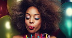Young African American woman with an afro celebrating her birthday blowing out candles on a cake at a party surrounded by colourful balloons and confetti. Young woman blowing out candles on a cake