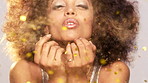Beautiful african american woman with an afro at a party blowing gold glitter and confetti from her hands.Happy young woman having fun blowing gold confetti and sparkles from her hands.