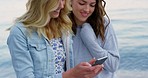 Two friends using a smartphone on the beach during their holiday. Cheerful women laughing and looking at a cellphone