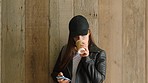 A young student posing in front of a wood wall using her smartphone and drinking a cup of coffee. A woman on her way to university drinking a cup of coffee and texting on her cellphone by a wood wall