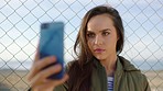 A happy young woman taking selfies on her mobile phone in front of a fence. A young woman smiling and standing in front of a fence to take selfies on her cellphone. A woman posing and taking selfies