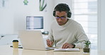 Mixed race creative entrepreneur using a laptop, writing notes in a notebook while listening to music on headphones. Young latino businessman and designer sitting alone and working from home office