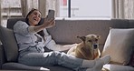 Happy young woman using her smartphone for a video call on the couch and waving at her house. Caucasian woman with her puppy. Fun and relaxing day on the couch with your pet and phone. Love and kiss human and animal.