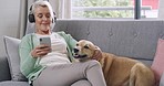 Mature caucasian woman dancing to music with headphones on cellphone and bonding with cute brown dog on a sofa at home. Carefree retired woman relaxing with pet while texting and browsing social media