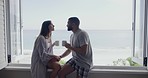Happy young interracial couple kissing while relaxing on window sill by the sea and enjoying their morning coffee. Young man and woman in pyjamas drinking tea having romantic moment