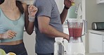 Two fit young mixed race people having fun while singing in a kitchen and making smoothies. Diverse couple being playful while making health shakes with fresh fruit and dancing in a bright apartment