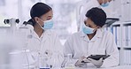 Two scientists using a microscope and digital tablet in a laboratory while doing sample analysis. A diverse female team of specialists wearing masks, working in an advanced lab, looking for a cure 