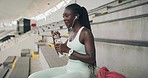 Young athlete listening to music, hydrating and getting ready to train at a stadium. Sporty black woman enjoying her favourite songs before a workout. Fit girl preparing to exercise at a sports club