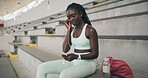 Trendy sports woman listening to music through earphones on her phone and sitting on stairs at the stadium. Young modern athlete taking a break from jogging and enjoying songs using latest technology