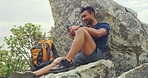 Fit active male using a phone, taking a break from hiking on a mountain while resting on a big rock. A smiling man browsing social media and texting on an app while relaxing outdoors in nature