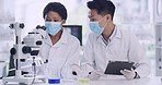 Two medical scientists use a tablet and microscope while checking a virus sample inside a research facility. Specialists enter data into a system and analyze it on a petri dish to develop a cure