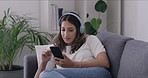 Young woman using phone to browse social media with headphones while relaxing on a sofa at home. Millennial streaming a podcast with free time on a sofa. Enjoying music and catching up with trends