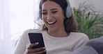 Young woman laughing while reading a text message on her phone and listening to music on wireless headphones. Beautiful female sitting alone in her living room and enjoying online streaming service