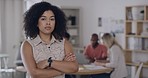 Portrait of a serious business woman standing with arms crossed in an office with her colleagues in the background. Young black entrepreneur looking focused and determined to lead her team to success