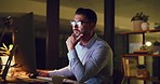 Young business man looking thoughtful while reading an email on a desktop computer and working late at night. One serious corporate male designer doing overtime to meet a deadline in a dark office