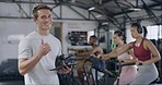 Fitness coach giving thumbs up and using a tablet while standing in a gym with his students in the background. Personal trainer smiling with his exercise class on air bikes going through a routine