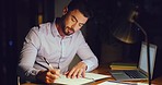 Young business man taking notes and writing in a diary while working on a laptop in an office late at night alone. One serious male professional reading an online search result and planning in a book