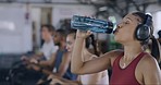 Sweaty female athlete drinking water inside a busy gym during her workout. Tired and thirsty Athletic woman hydrating and refreshing while exercising. Active fit lady resting in a fitness facility