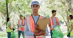 Portrait of female volunteer coordinator looking serious while holding a clipboard. Group of diverse people volunteering and doing charity work in a local park. Young people doing a cleanup outdoors