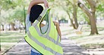 Rearview of a woman putting on a reflective safety vest representing authority while standing outside in a park. Female public servant or volunteer working outdoors on a bright and sunny summer day