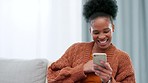 Happy woman scrolling and browsing through social media online with her phone while sitting on the couch at home alone. One cheerful black female typing and sending a text message to her friend