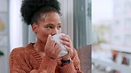 Woman drinking a cup of coffee and looking at the view from her window in the lounge at home. One happy, comfy, and peaceful black female smiling while relaxing and enjoying a warm mug of tea alone