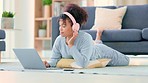 Young African American woman lying on her lounge floor chatting online. Female barefoot with blue tracksuit and pink headphones smiles typing on laptop. Searching for music and listening to songs