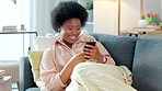 African American woman on sofa laughing using phone. Lady typing and smiling in a comfortable environment. Adult having a conversation through text and replying to a message.