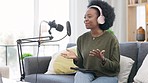 Young woman producing a podcast while using a digital tablet for research on the couch in the lounge at home alone. Happy black female entrepreneur talking on a web show from the comfort of her sofa