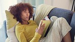 Beautiful afro woman reading intriguing book while relaxing on the couch at home. Content and smart student studying literature and reading an educational book in her free time in her cozy apartment
