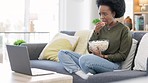 African woman watching a horror movie on a laptop and eating popcorn while sitting on the couch at home. Terrified black afro female enjoying her online subscription with a variety of scary films