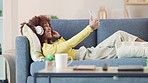 Trendy young black woman wearing headphones and taking selfies while relaxing on a couch at home. African American female enjoying modern apps and technology while streaming online music on a weekend